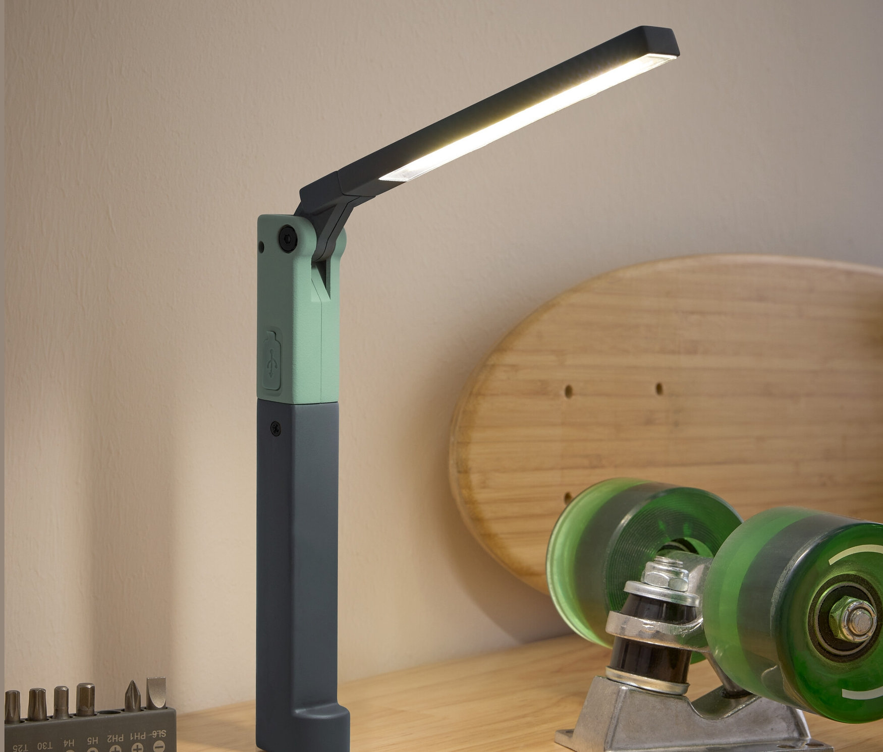 Na drevenom stole stojí LED pracovná lampa, skrutkovač, sada bitov a skateboardové príslušenstvo.