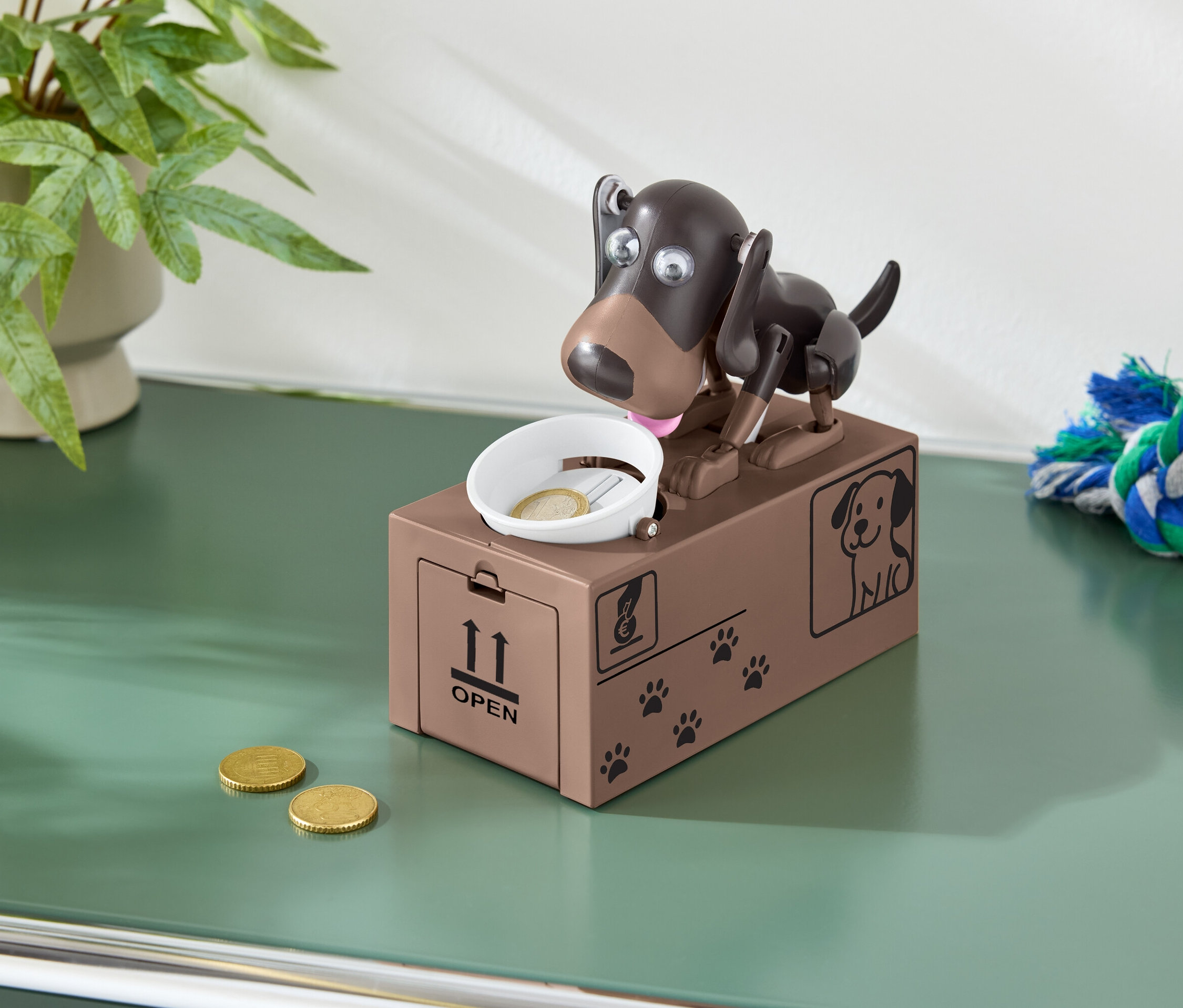 Hnedá pokladnička v tvare psa s mincou v miske a dvoma mincami na zelenom stole.