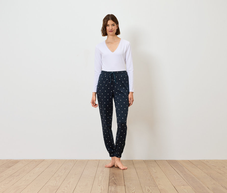 Žena stojí a má na sebe biele tričko s dlhým rukávom a tmavomodré nohavice so vzorom.