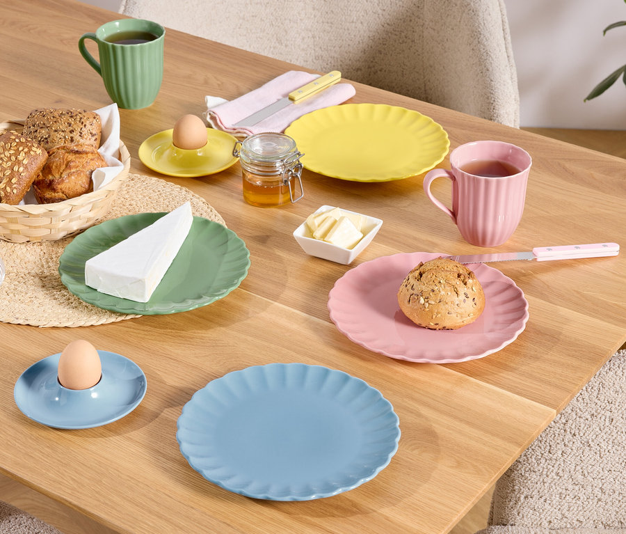 Na drevenom stole sú prestreté farebné taniere, šálky a misky s jedlom, vrátane chleba v košíku, vajíčka v kalíšku a kúska syra.