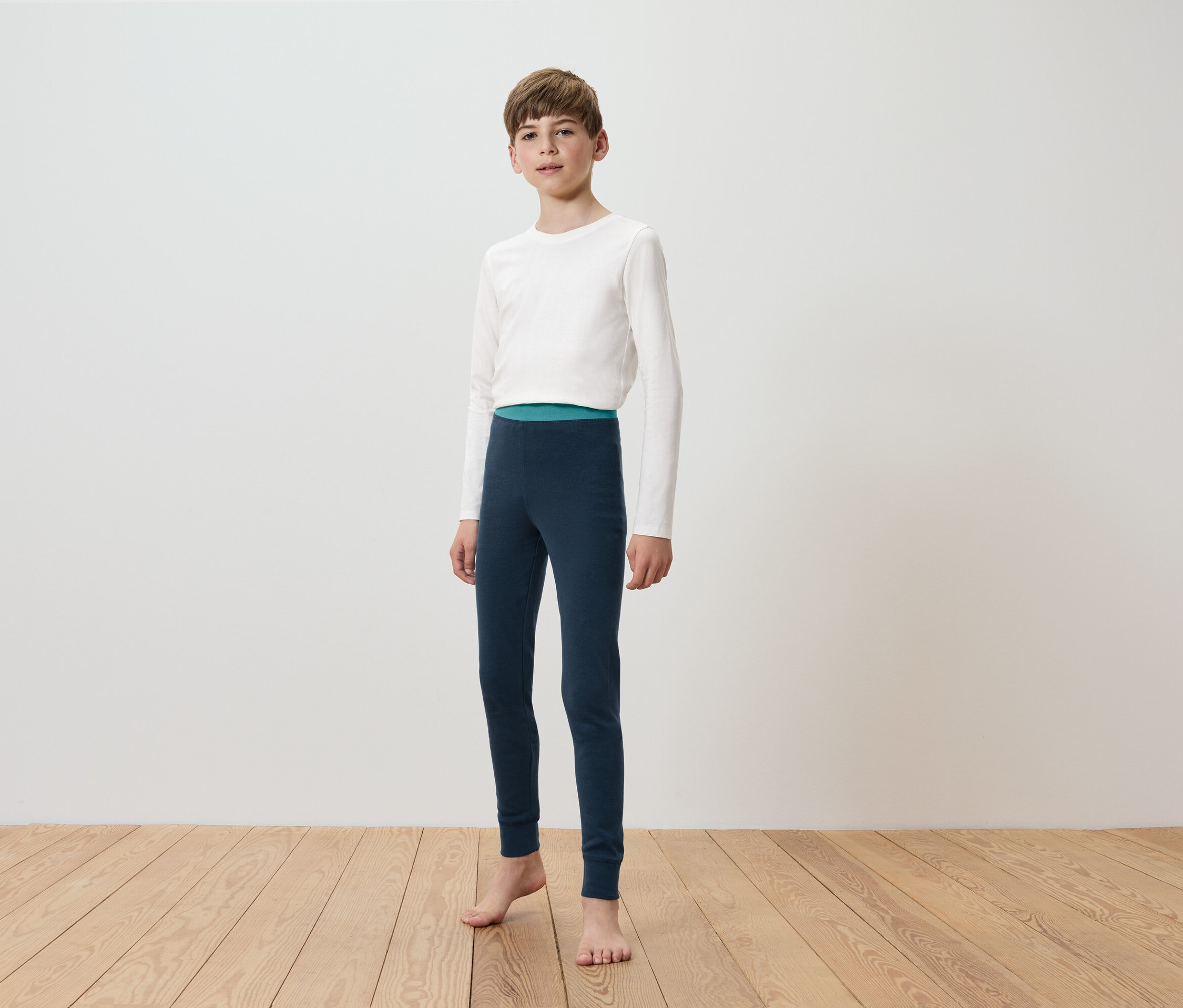Chlapec v bielom tričku s dlhým rukávom a tmavomodrých legínach stojí na drevenej podlahe pred bielou stenou.