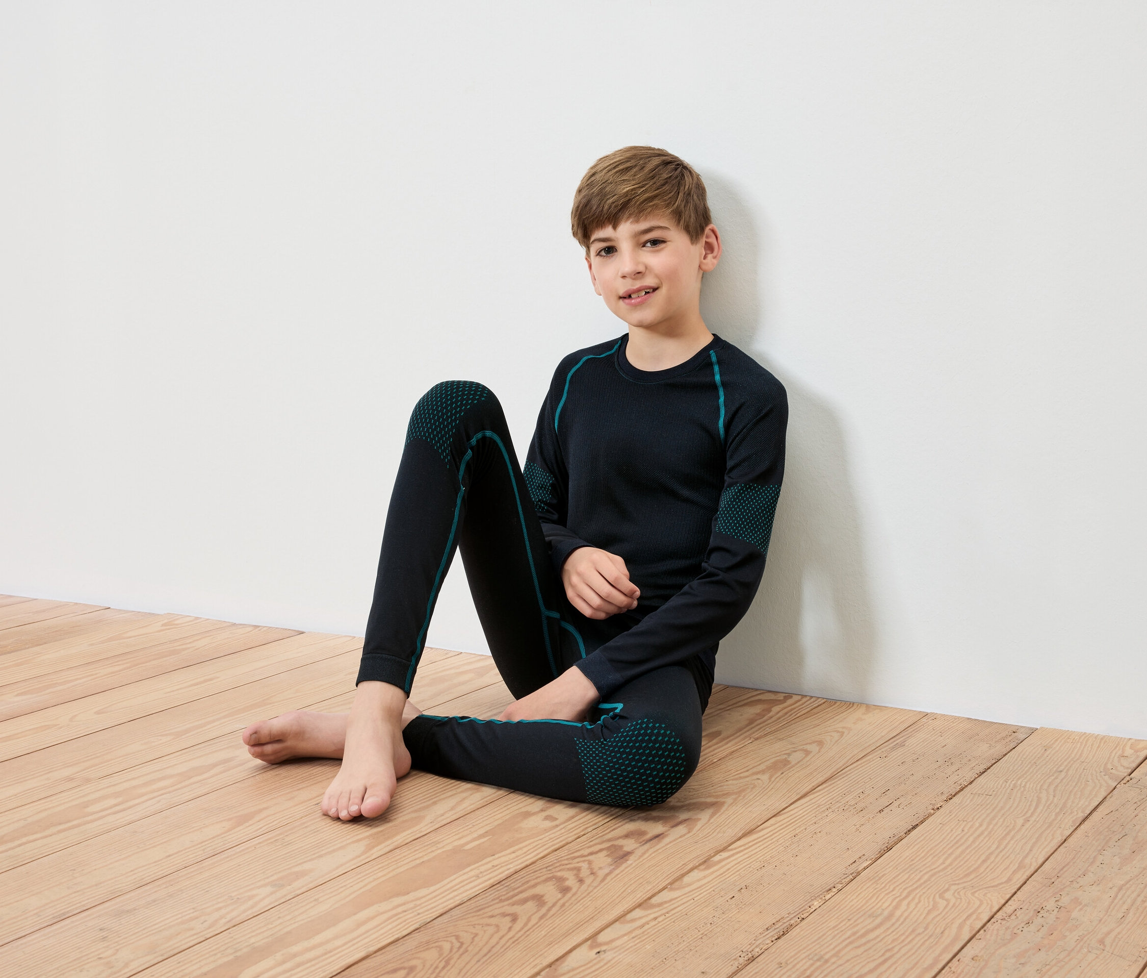 Chlapec sedí na drevenej podlahe opretý o bielu stenu, má na sebe čierne termooblečenie s tyrkysovými detailmi.