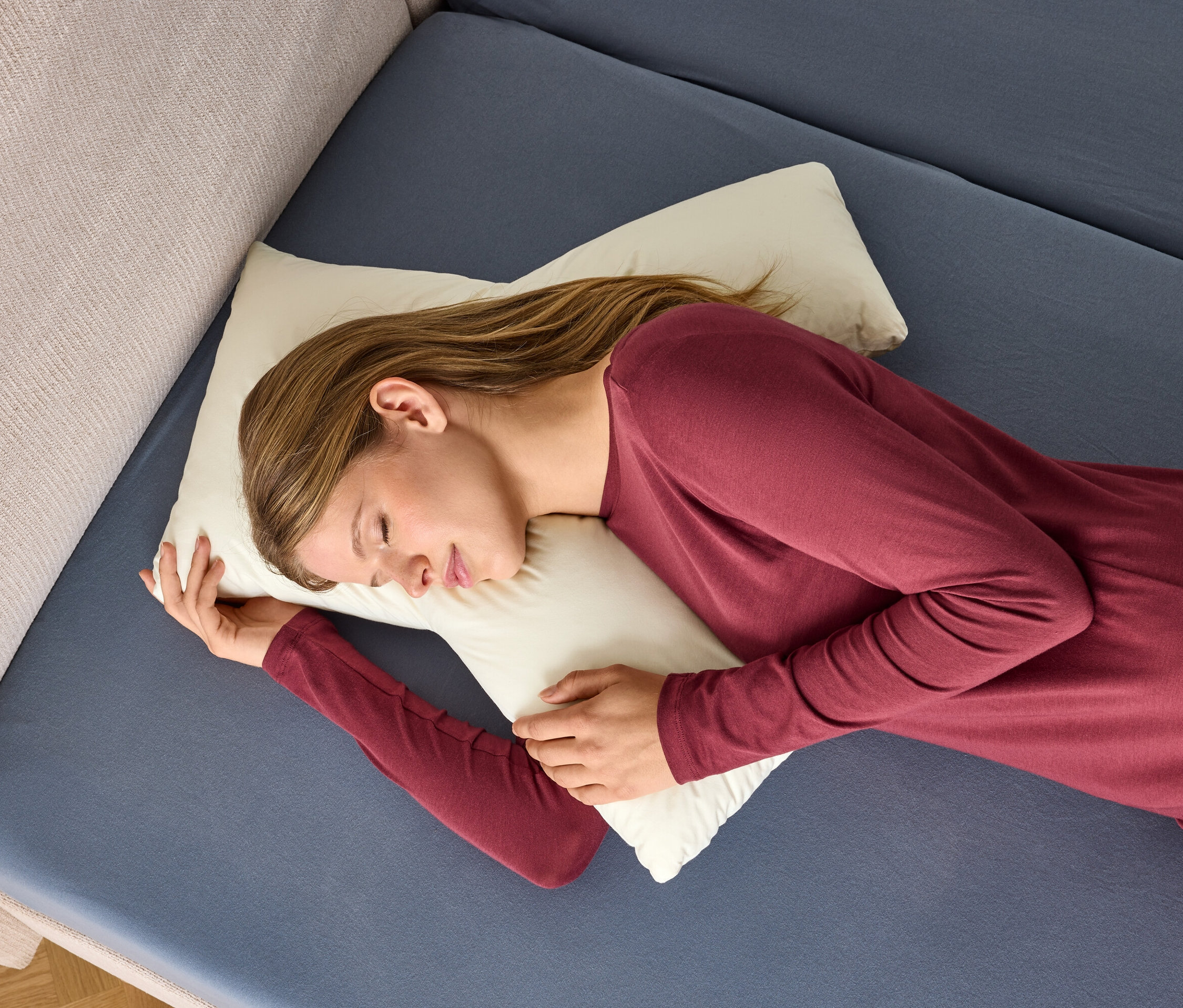 Žena v bordovej nočnej košeli spí na bielom vankúši na modrej posteli.