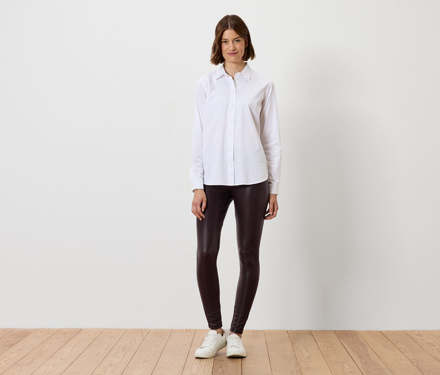 Žena v bielej košeli a hnedých legínach stojí na drevenej podlahe.