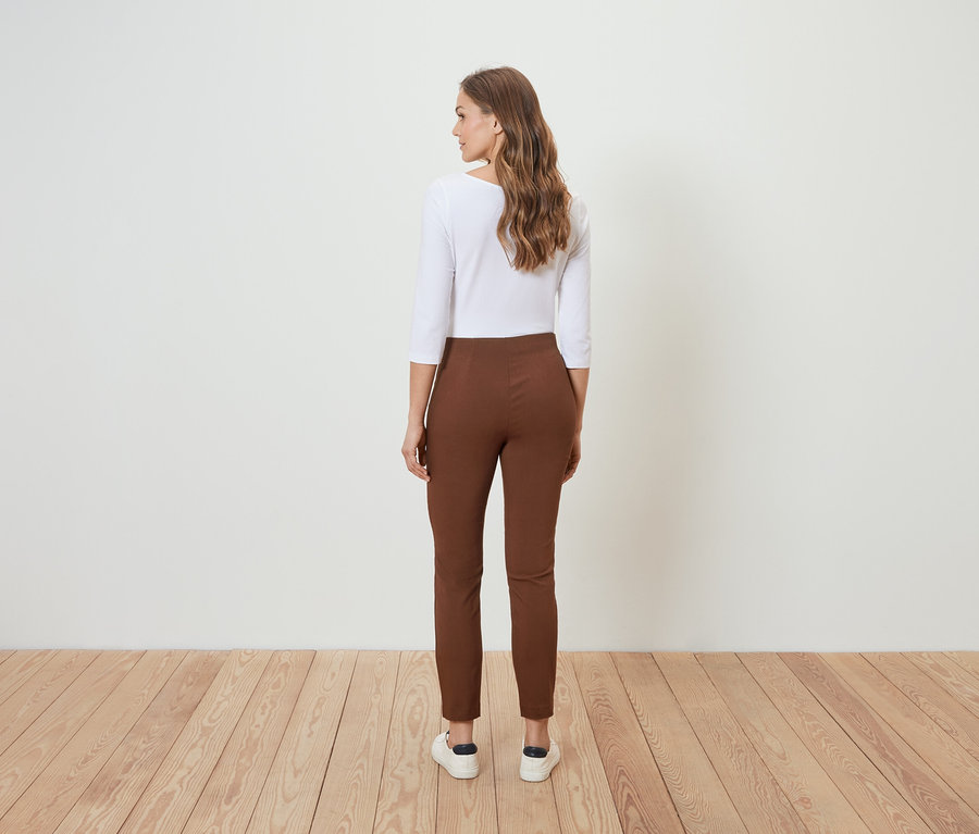 Žena stojí chrbtom k fotoaparátu a má na sebe hnedé nohavice a bielu blúzku.