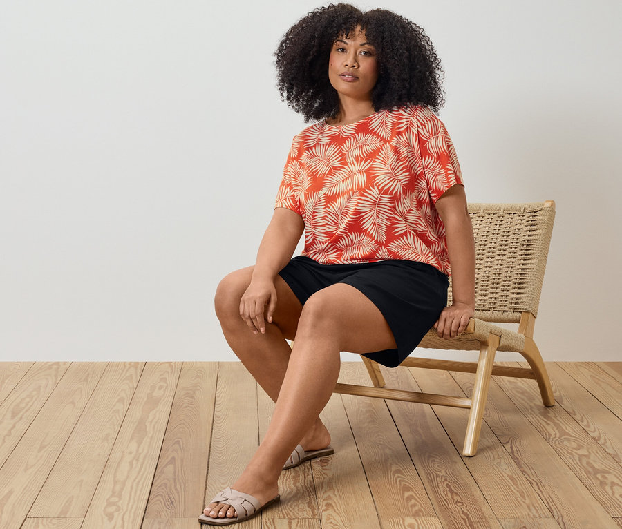 Žena sedí na stoličke a má na sebe vzorované blúzkové tričko a čierne džersejové šortky.