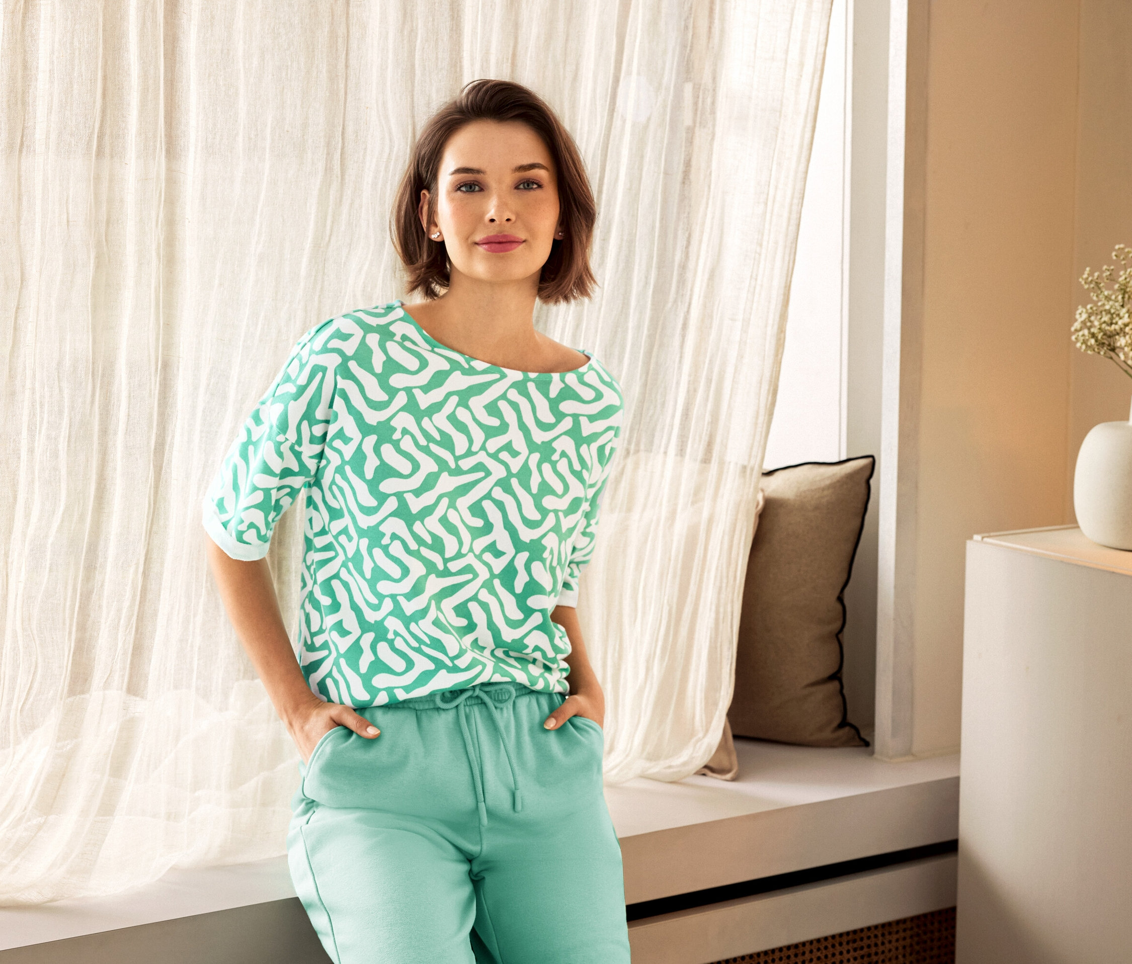 Žena v zelených nohaviciach a vzorovanej blúzke pózuje vedľa okna. V pozadí je vidieť váza.