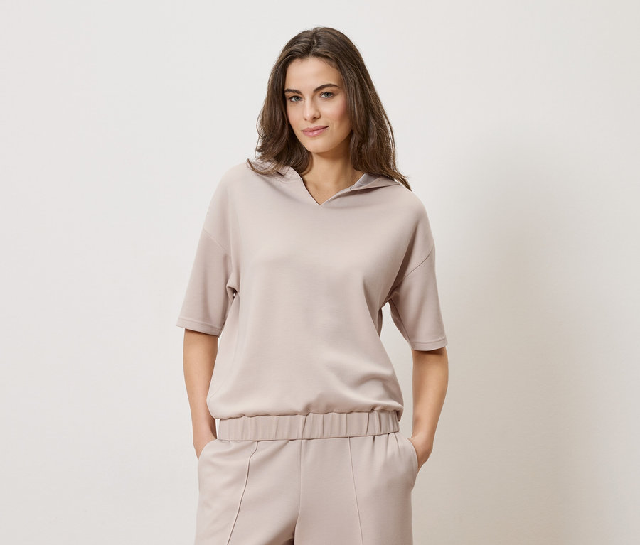 Žena s hnedými vlasmi má na sebe béžovú mikinu s kapucňou a zodpovedajúce nohavice na jednoduchom pozadí.