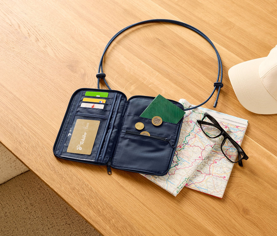 Otvorená, tmavomodrá cestovná peňaženka s kartami, pasom a mincami na drevenom stole, spolu s mapou, čiernymi okuliarmi a bielou šiltovkou.