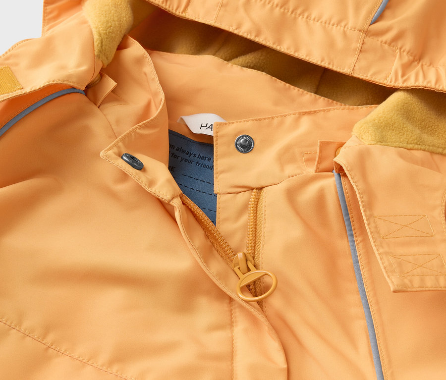 Detail oranžovej bundy s kapucňou, žltou flísovou podšívkou, zipsom a patentkami.