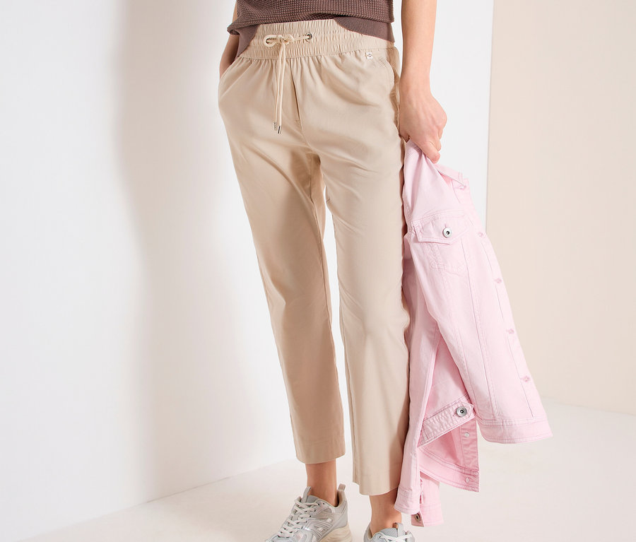 Žena v pohodlných nohaviciach CECIL »Tracey«, hnedom tope a strieborných teniskách. V ruke drží ružovú bundu.