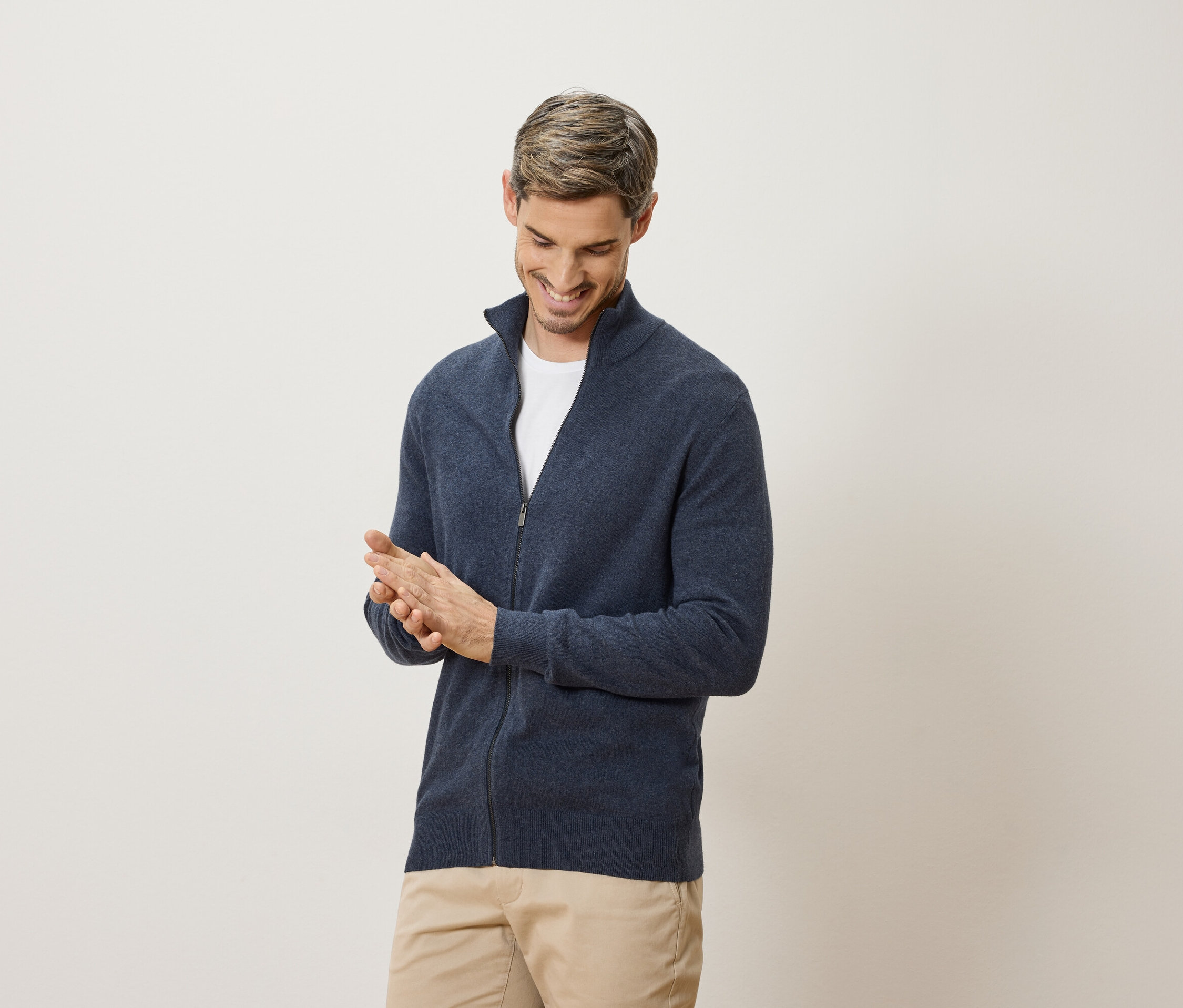Muž sa usmieva a trie si ruky. Má na sebe tmavomodrý kardigán a béžové nohavice.