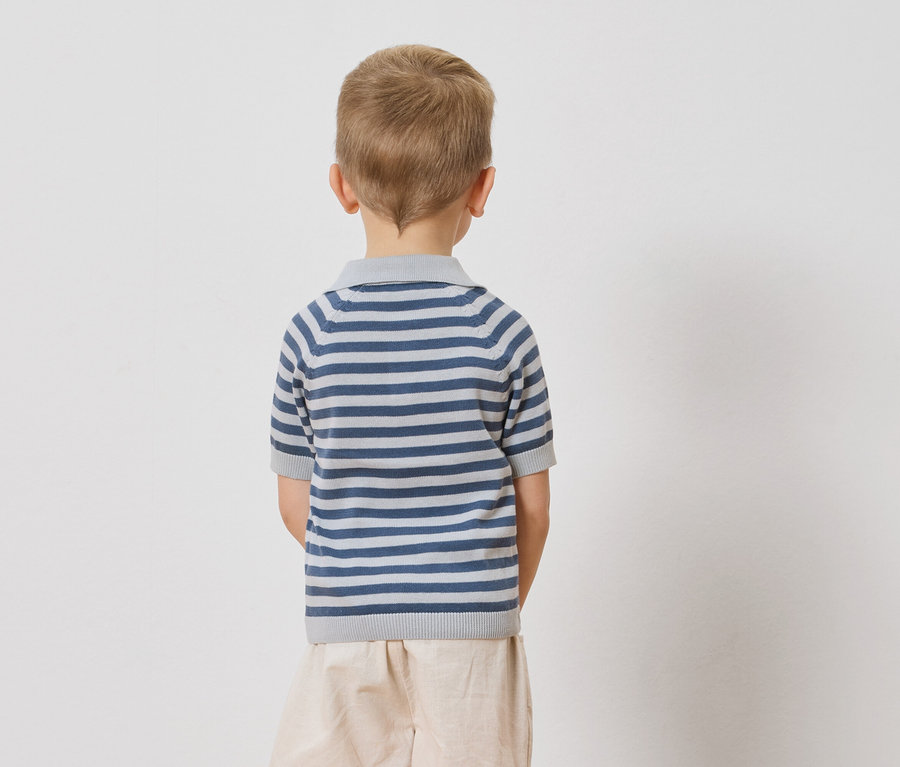 Pohľad zozadu na dieťa s blond vlasmi v modro-bielo pruhovanom polo tričku a béžových šortkách na bielom pozadí.