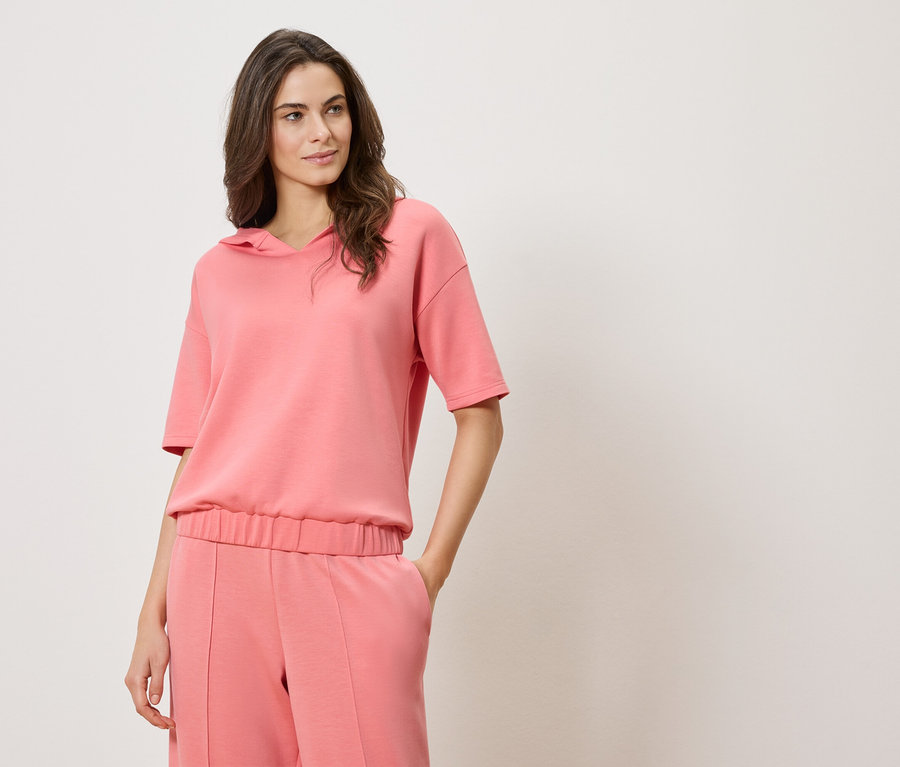 Žena má na sebe ružovú blúzu s krátkym rukávom a kapucňou a ružové nohavice.