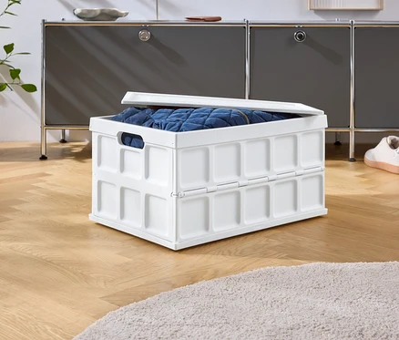 Na drevenej podlahe stojí biely skladací box s modrým obsahom, za ním sivá skrinka a topánky.