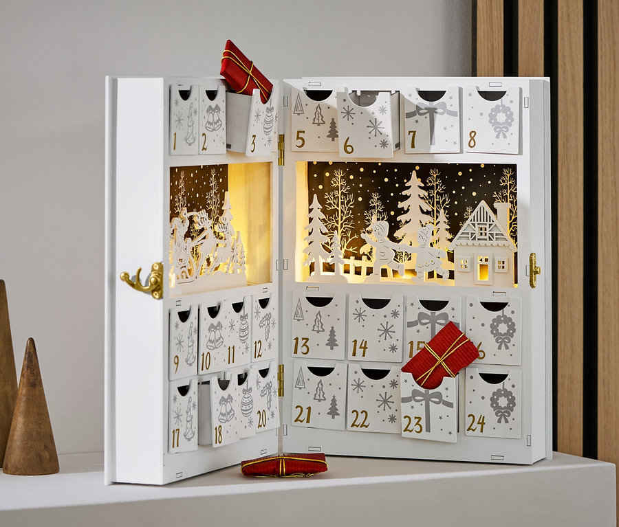 Otvorený biely adventný kalendár s ozdobami a darčekmi, vedľa troch drevených kužeľov.