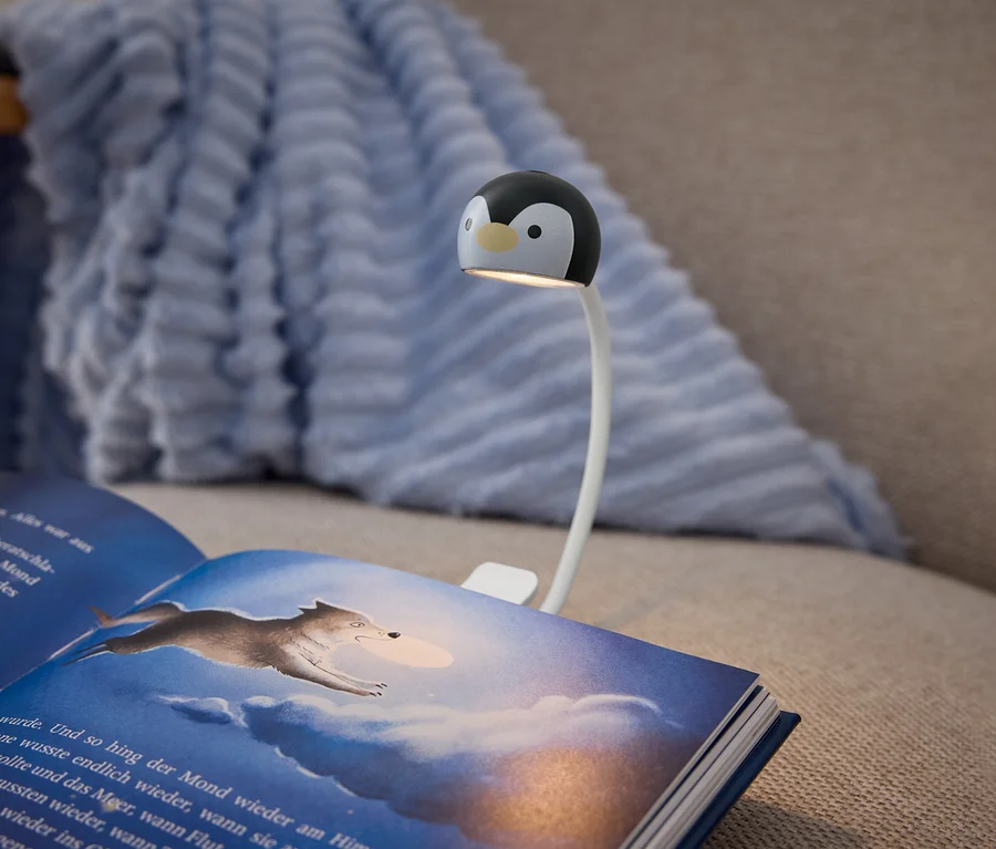 Otvorená kniha s tmavomodrým obalom leží na béžovej pohovke, osvetlená klipsovou lampou. Lampa má biele flexibilné rameno a čierno-bielu hlavu v tvare tučniaka, ktorá vrhá svetlo na stránky knihy. Pravá strana zobrazuje ilustráciu vlka skákajúceho cez zamračenú, modrú oblohu. Svetlomodrá, textúrovaná deka je prehodená cez operadlo pohovky v pozadí.