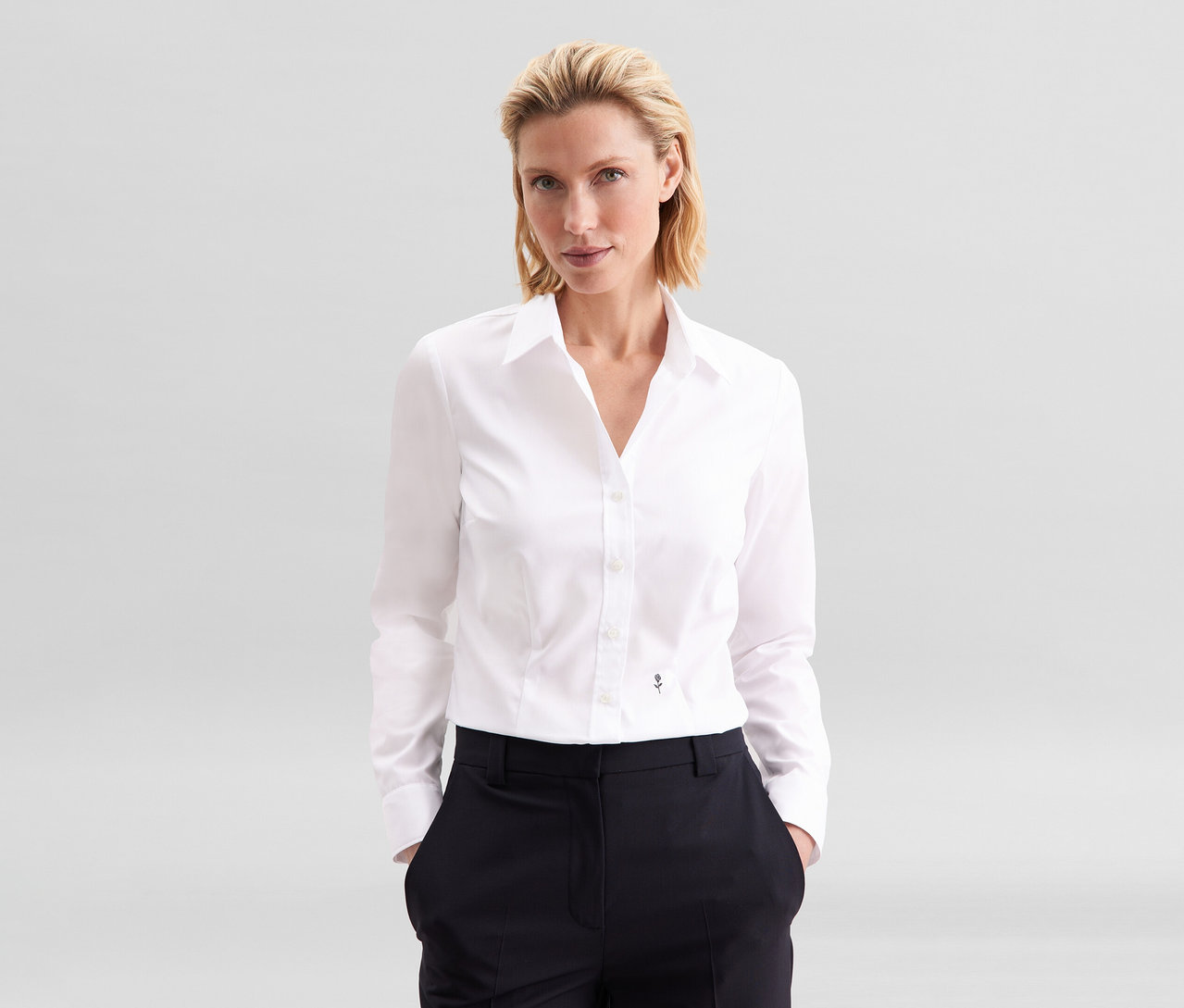 Blondínka stojí s rukami vo vreckách, oblečená v bielej blúzke Seidensticker Slim Fit non iron a tmavých nohaviciach.