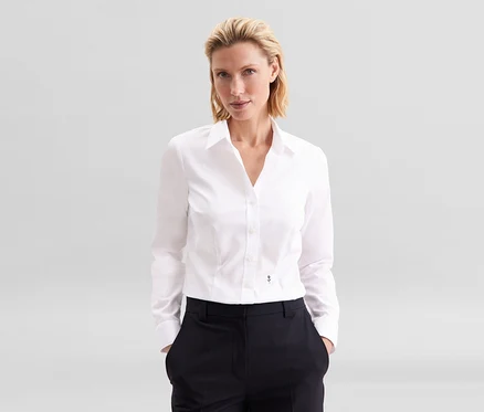 Blondínka stojí s rukami vo vreckách, oblečená v bielej blúzke Seidensticker Slim Fit non iron a tmavých nohaviciach.