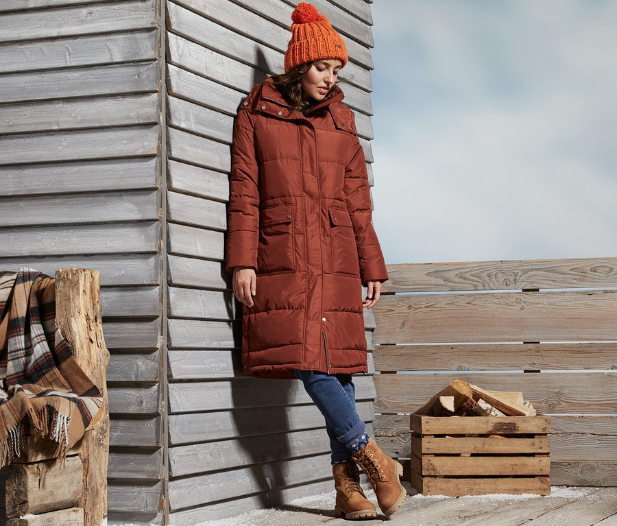 Žena opretá o drevenú stenu má na sebe prešívaný kabát s kapucňou, rifle a hnedé topánky.