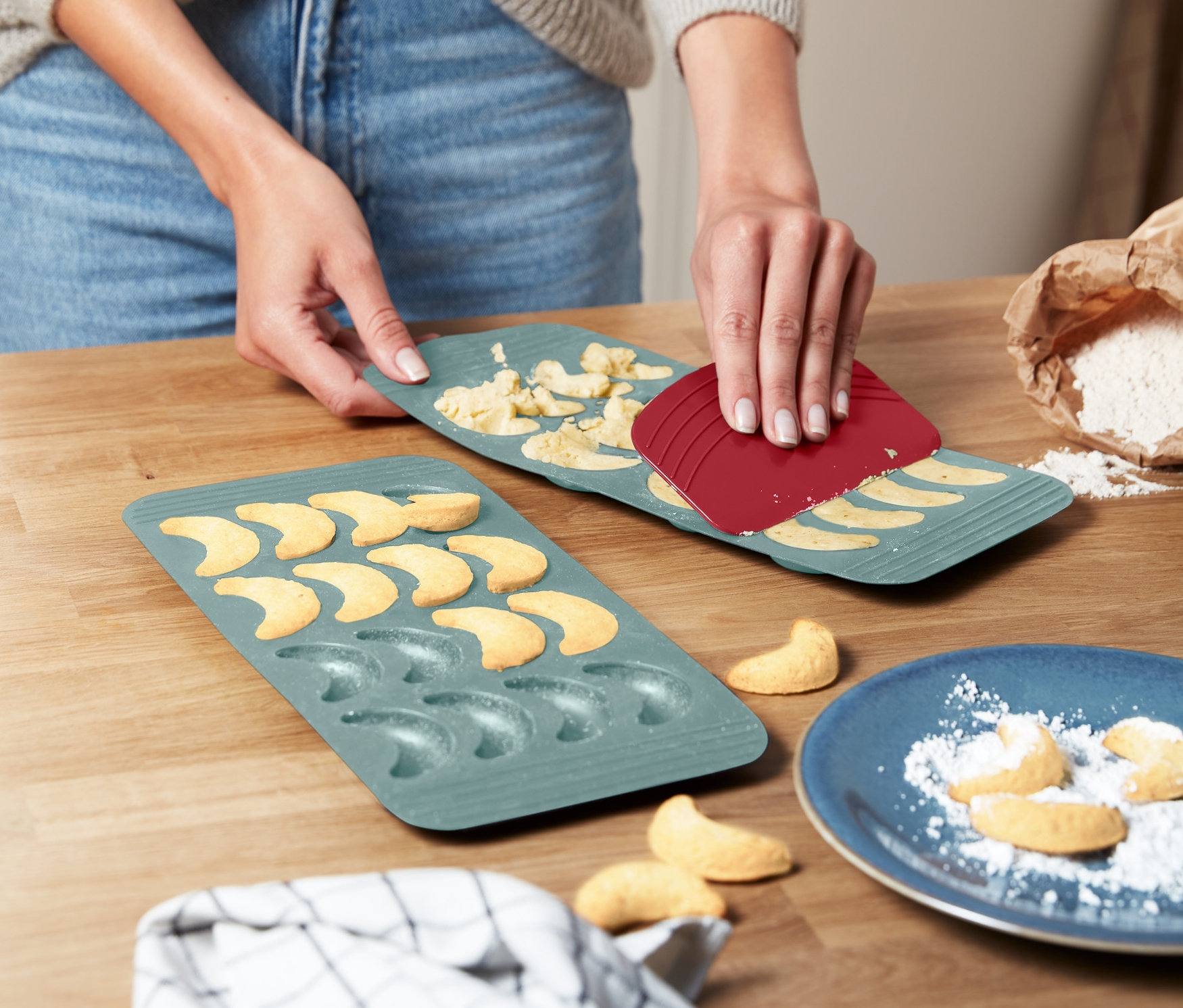 Žena pomocou červenej špachtle oddeľuje cesto z dvoch zelených foriem na pečenie vanilkových rožkov.