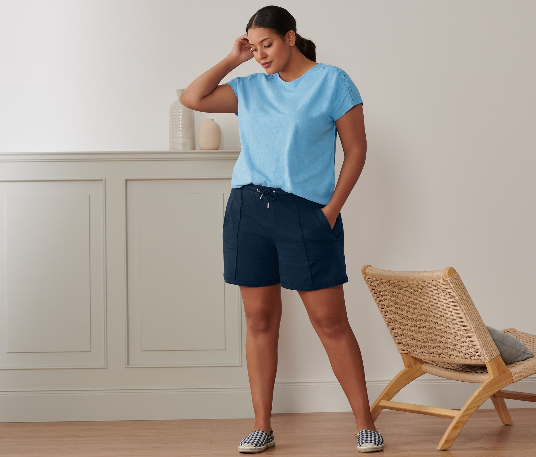 Žena stojí v miestnosti a má na sebe svetlomodré tričko z ľanovej zmesi, teplákové šortky a espadrilky.