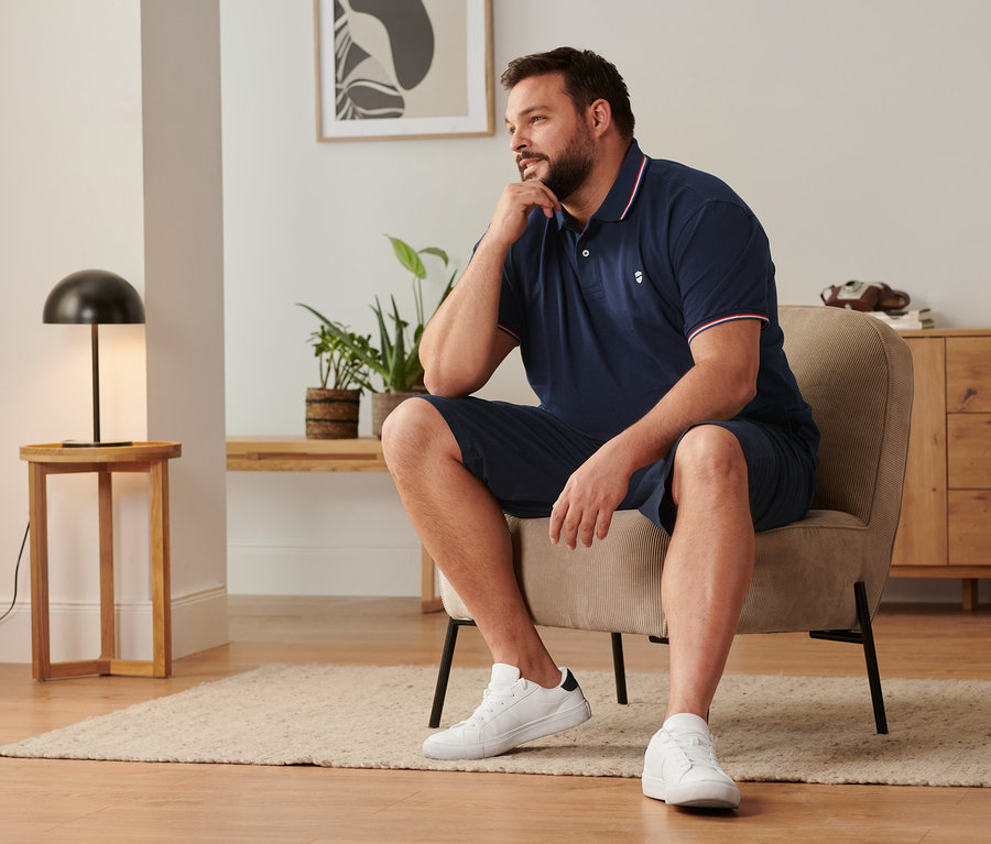 Muž sedí na stoličke, má na sebe námornícke modré šortky chino, topánky sneaker a piké polokošeľu v námorníckej modrej farbe.