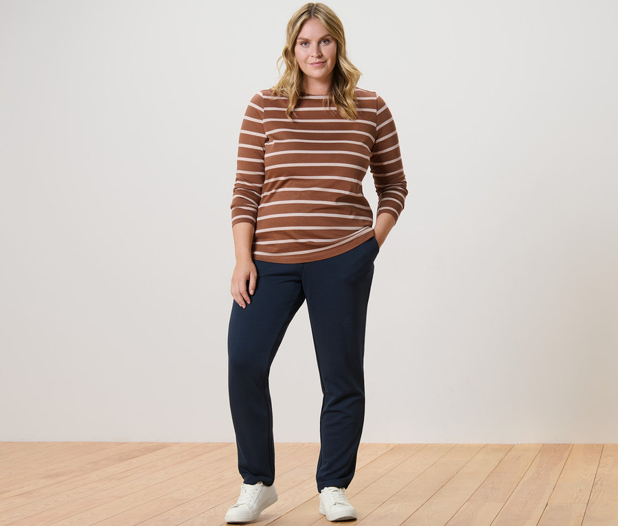 Žena pózuje v prúžkovanom tričku s dlhými rukávmi v koňakovej farbe a v teplákoch.
