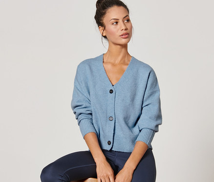 Žena sedí na stoličke a má na sebe modrý kardigán z hrubej pleteniny.
