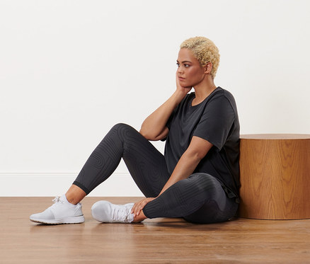 Žena sedí na podlahe v antracitovom športovom tričku a čiernych obojstranných športových legínach.