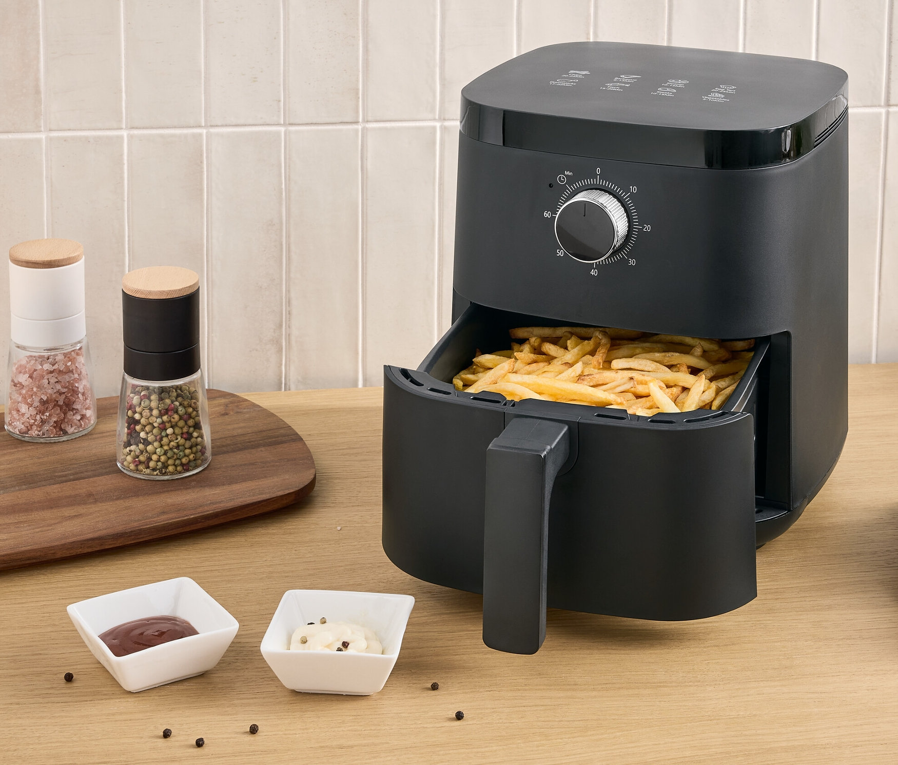 Čierna teplovzdušná fritéza s hranolčekmi vo výsuvnej zásuvke stojí na stole vedľa taniera s hranolčekmi a dvoch omáčok.