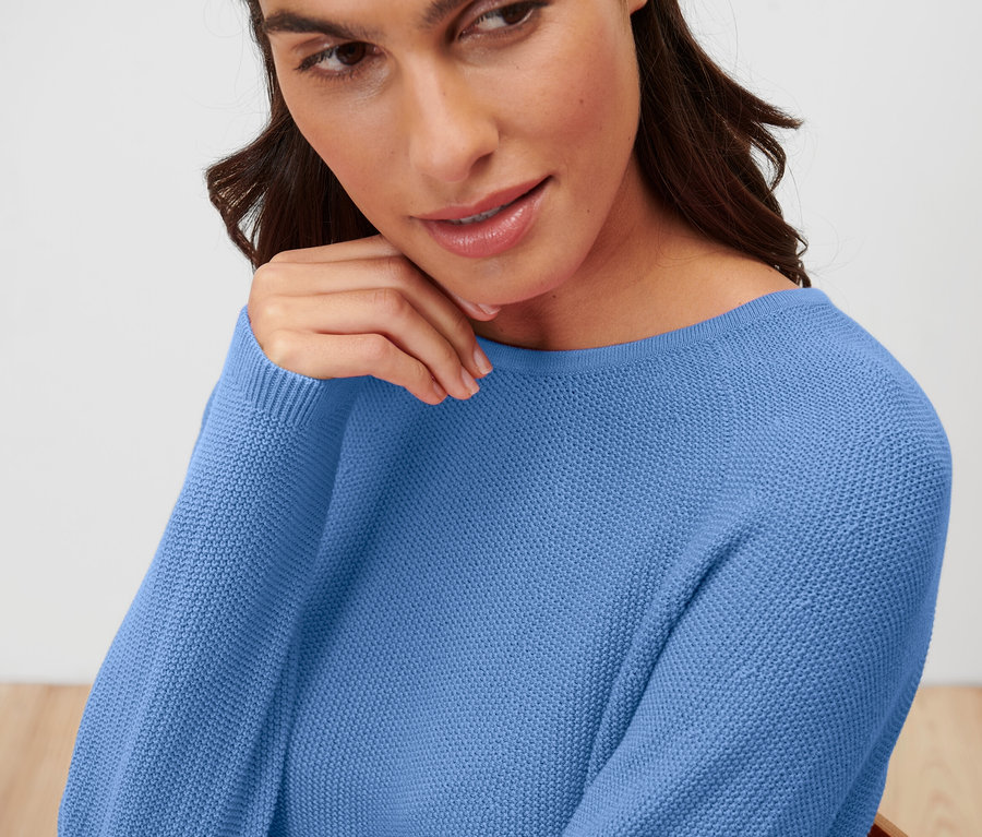 Žena s hnedými vlasmi nosí modrý pletený pulóver.