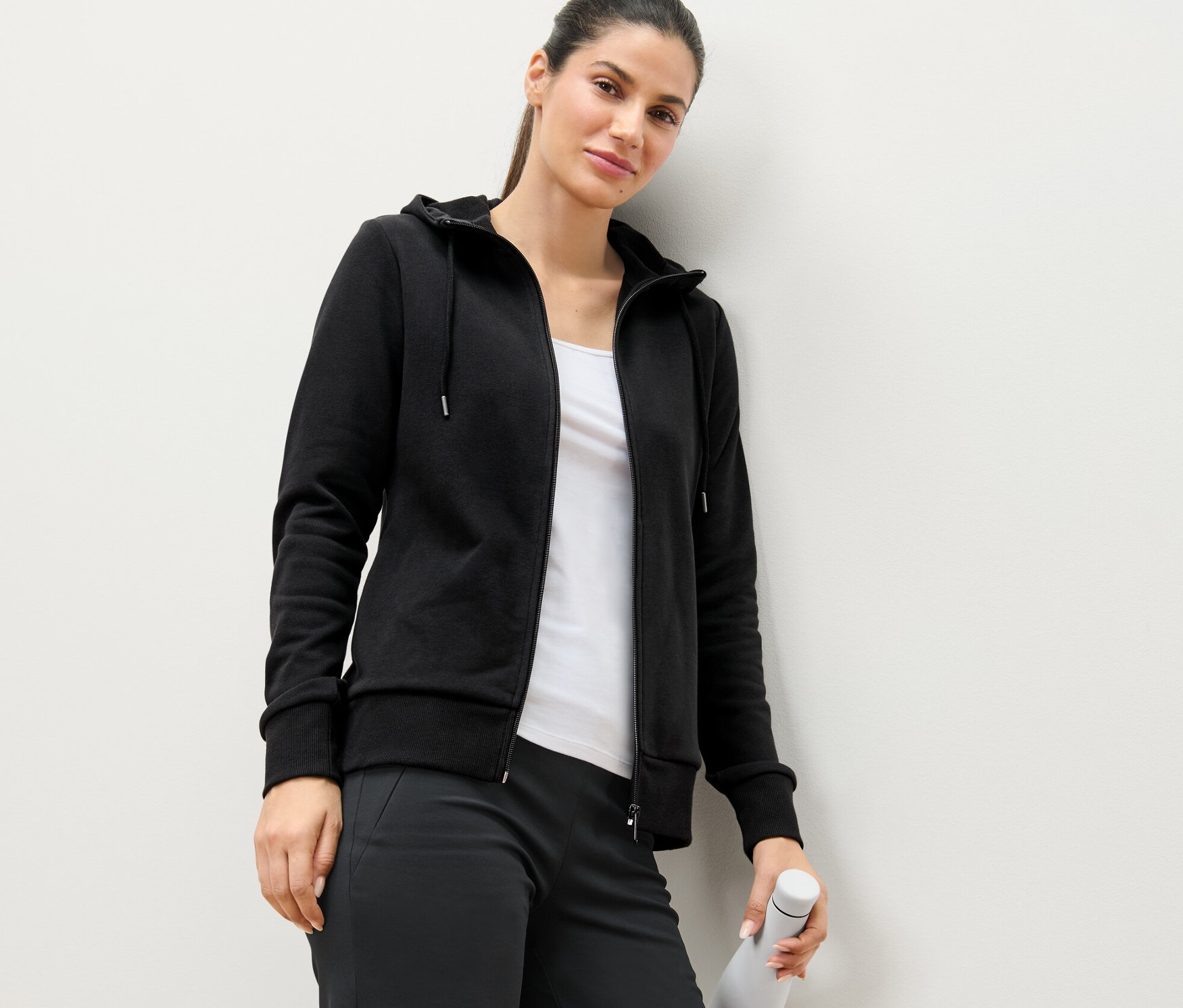 Žena opretá o stenu má na sebe čiernu mikinu a drží bielu fľašu s vodou.