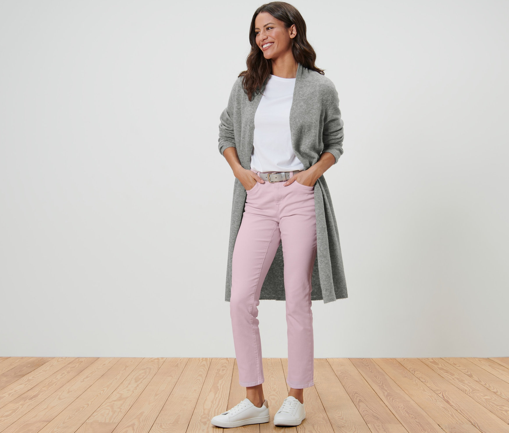 Žena v dlhom kardigáne bez zapínania, bielom tričku, ružových nohaviciach a bielych teniskách stojí na drevenej podlahe.