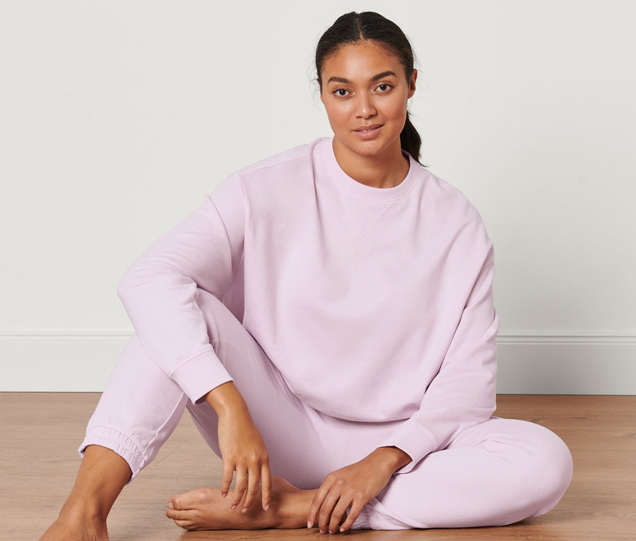 Žena sedí na podlahe a má na sebe svetloružovú oversized mikinu na jogu.
