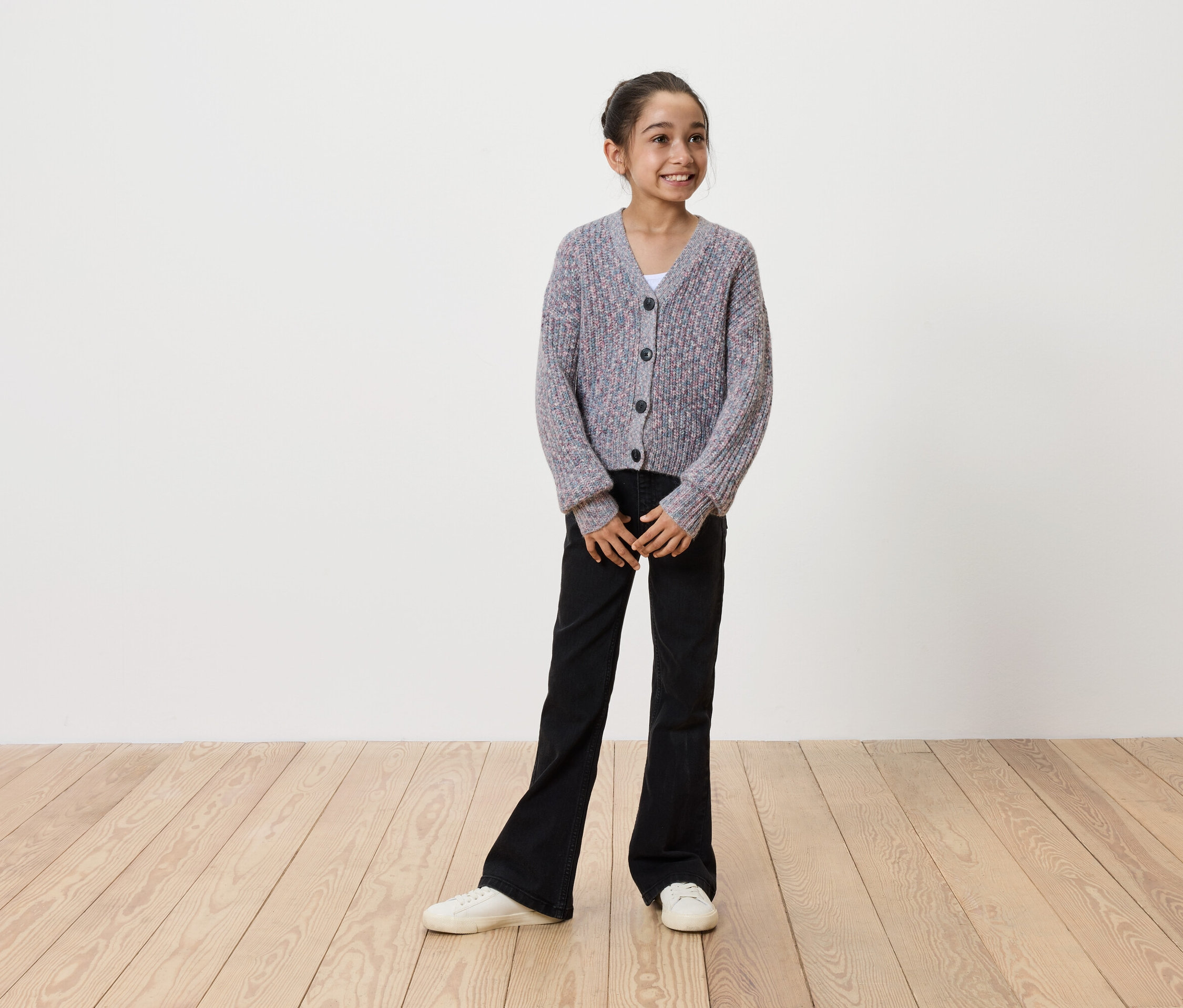 Usmievajúce sa dievča v sivom pletenom kardigáne na gombíky, čiernych zvonových nohaviciach a bielych teniskách stojí na drevenej podlahe pred bielou stenou.