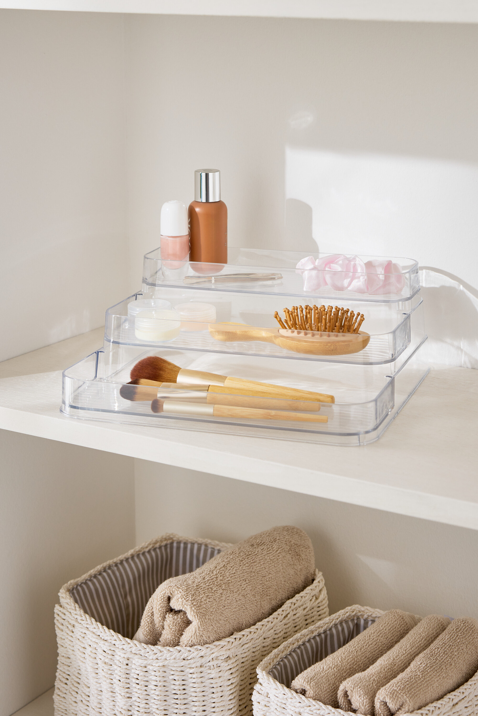 Trojposchodový priehľadný organizér na líčidlá na bielej polici vedľa košíkov s uterákmi.