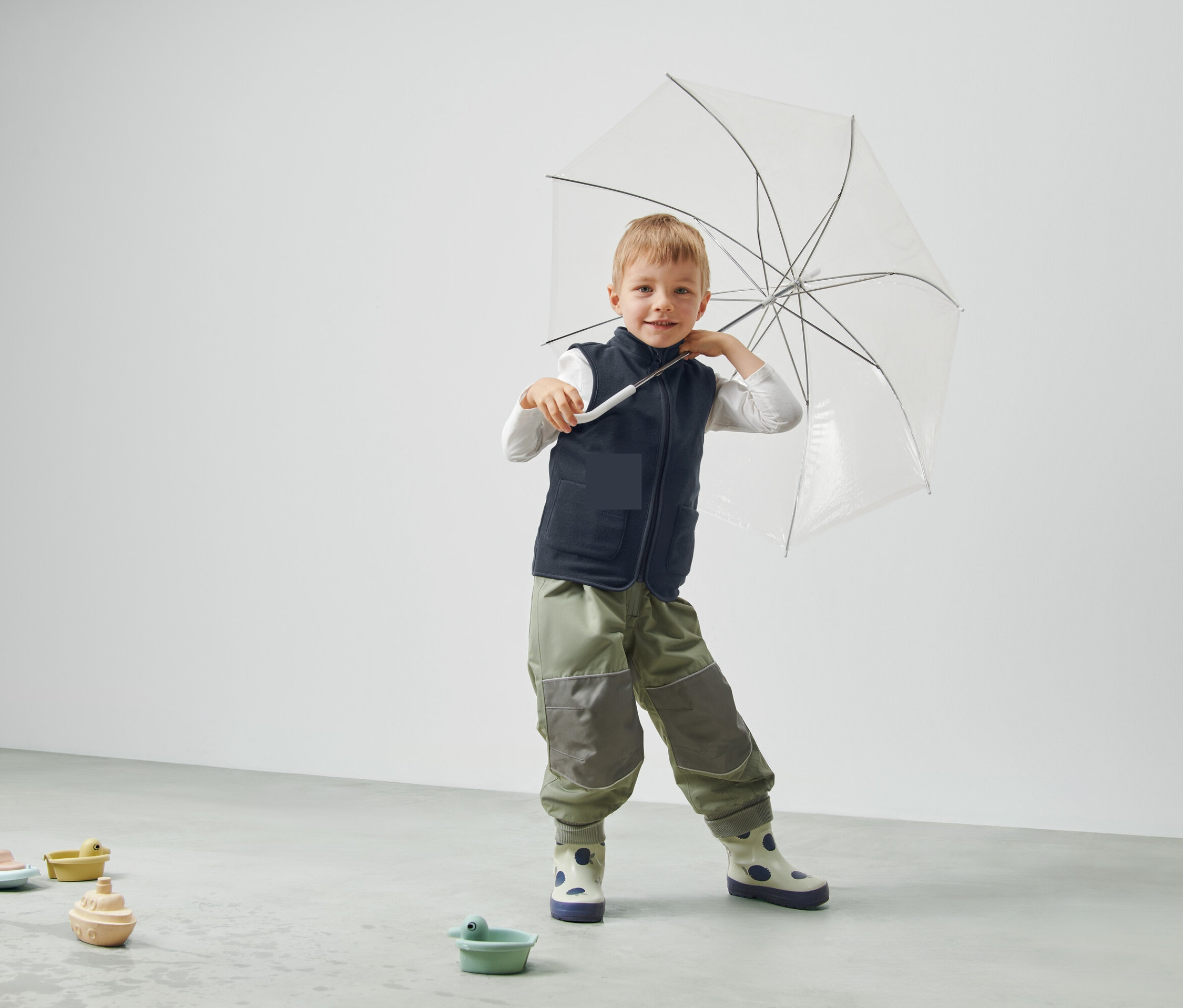 Chlapec drží priehľadný dáždnik, má na sebe vestu a nohavice a stojí v miestnosti.