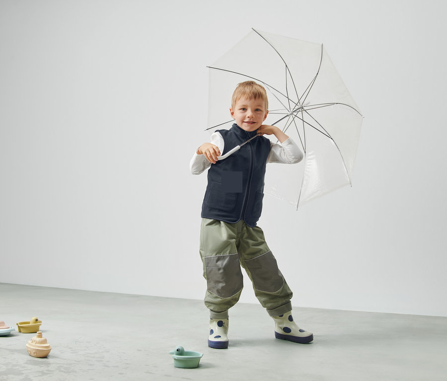 Chlapec drží priehľadný dáždnik, má na sebe vestu a nohavice a stojí v miestnosti.