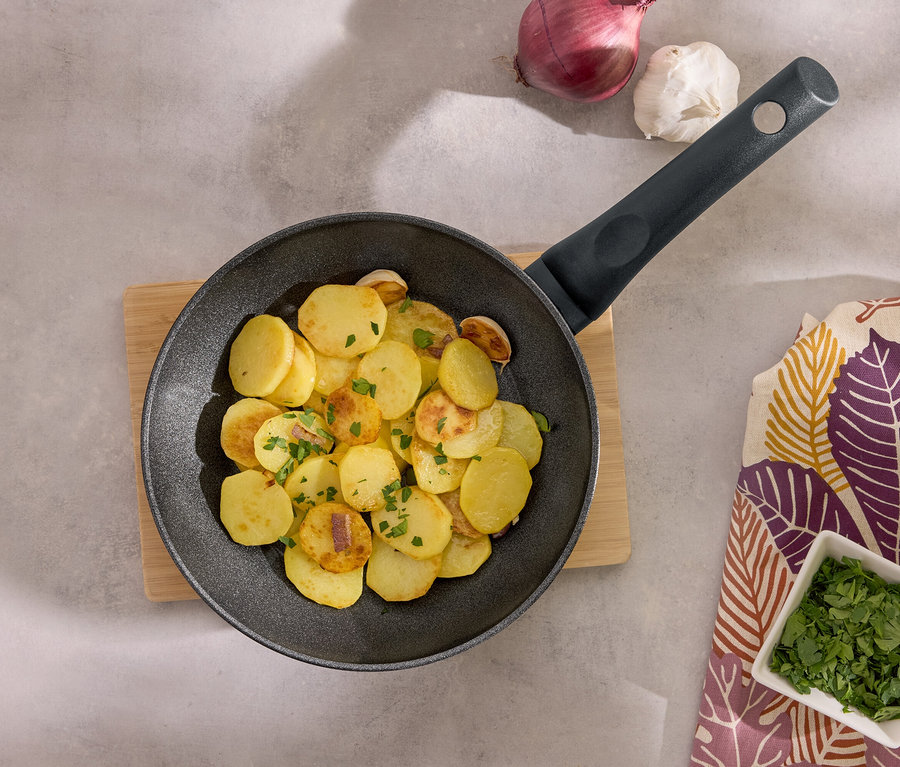 Vyprážané zemiaky na čiernej panvici na drevenej doske s cesnakom, cibuľou a petržlenovou vňaťou.