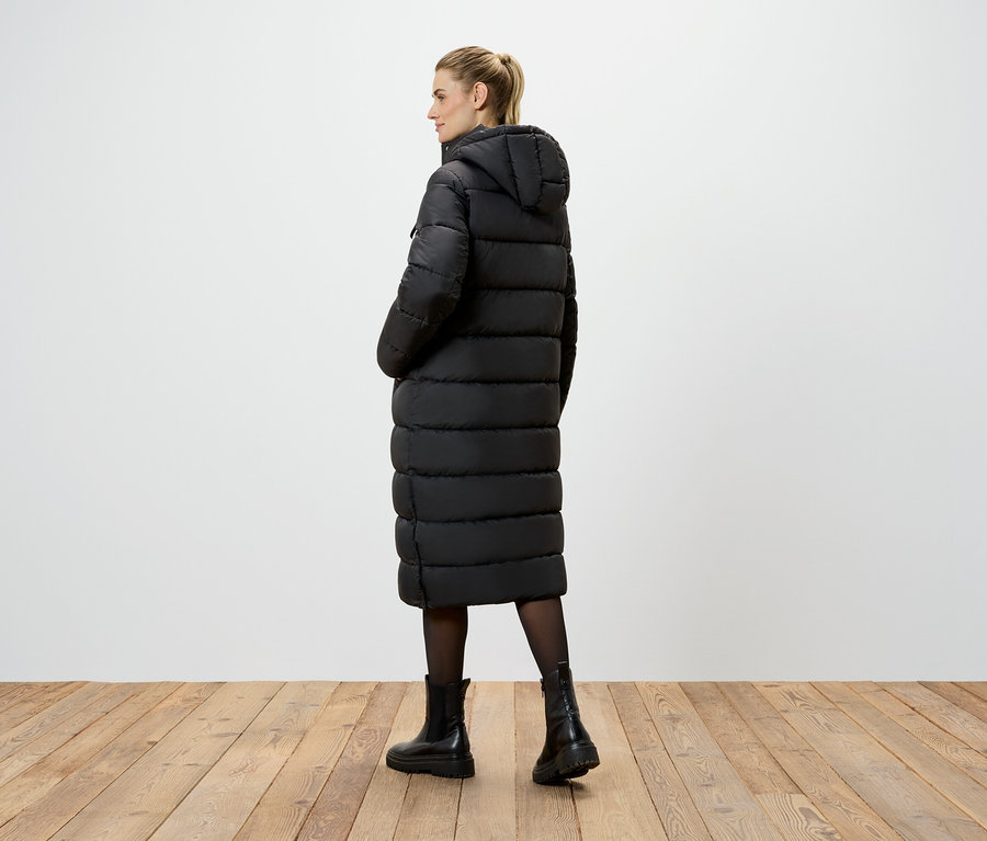 Žena stojí chrbtom ku kamere a má na sebe dlhý čierny kabát s kapucňou, čierne pančuchy a čierne topánky.