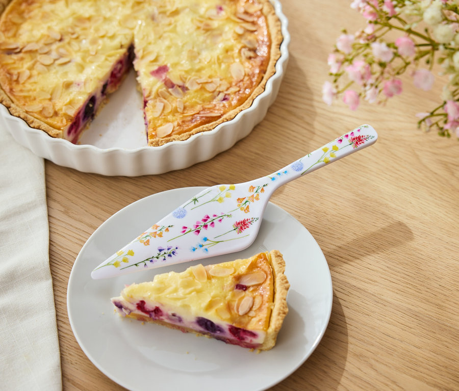 Koláč s ovocím a mandľami v bielej forme, kúsok koláča na tanieri a kvetinový servírovací nôž.