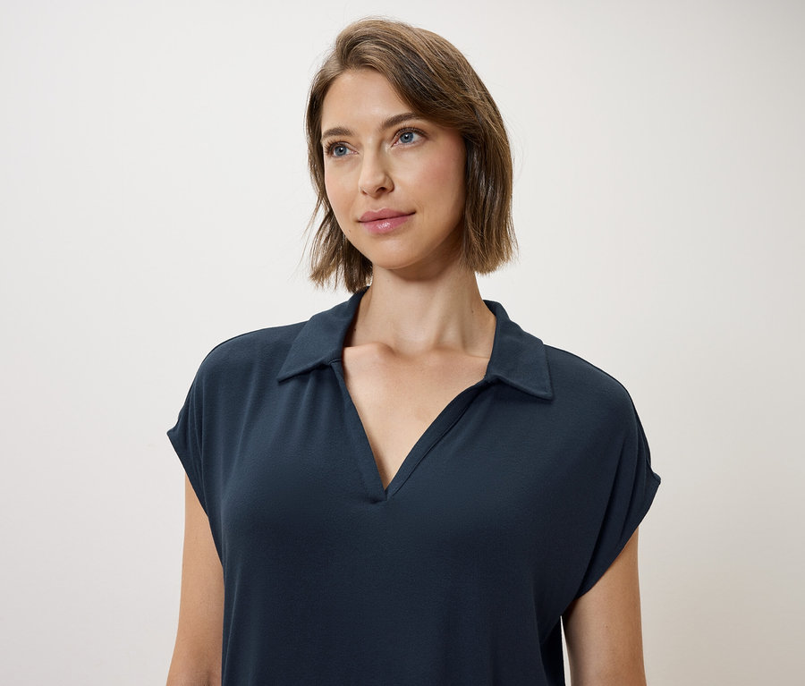 Žena s hnedými vlasmi a modrými očami má na sebe tmavomodré polokošeľové šaty.