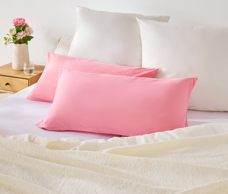 Na posteli sú dva ružové a dva biele vankúše. Na nočnom stolíku je váza s kvetmi.