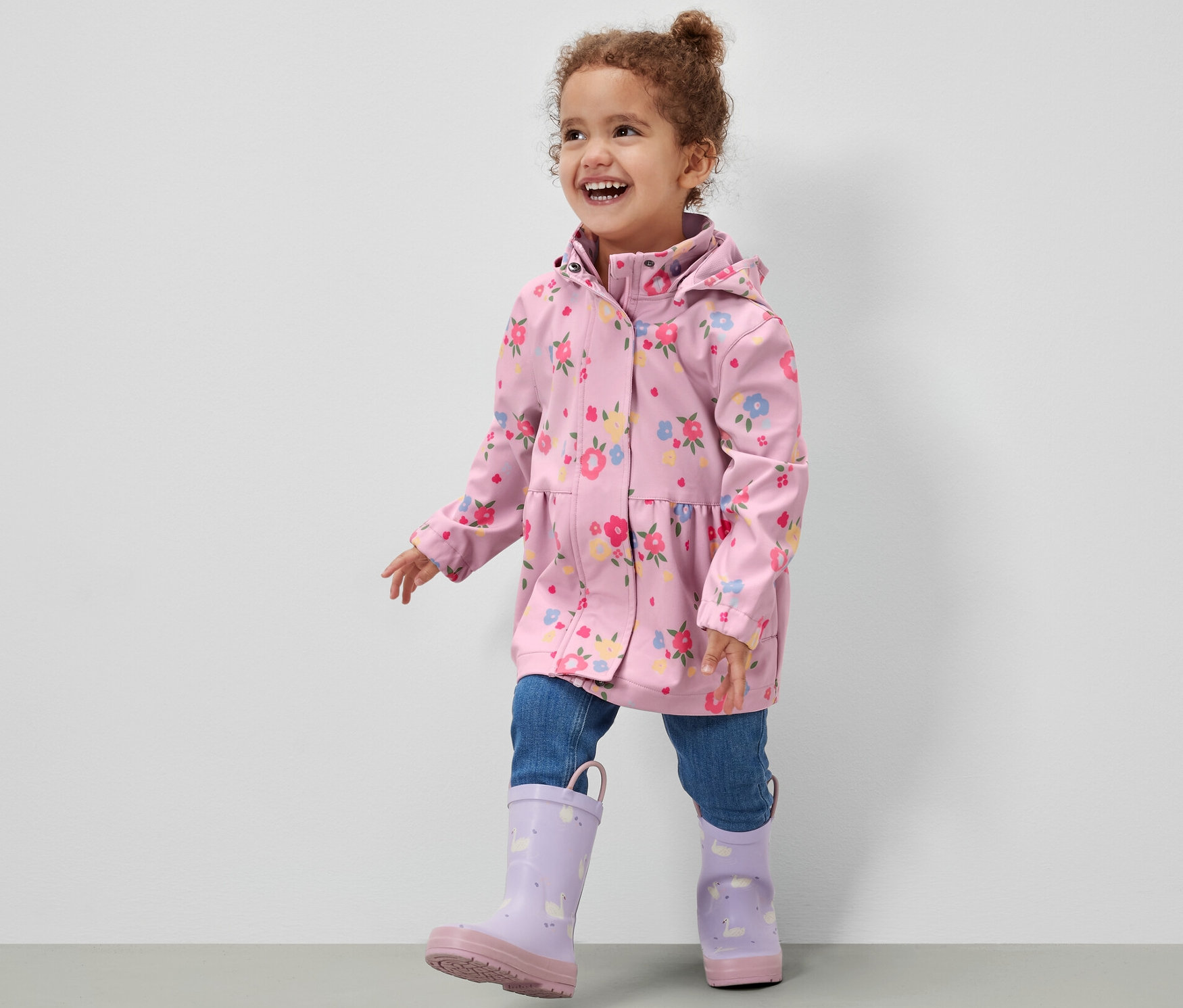 Usmievajúce sa dievčatko v detskej softshellovej bunde s kvetinovým vzorom, džínsoch a fialových gumákoch kráča po sivej podlahe pred bielou stenou.