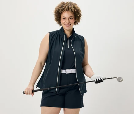 Žena drží golfovú palicu, má na sebe tmavomodrý funkčný top, softshellové funkčné šortky a softshellovú vestu.