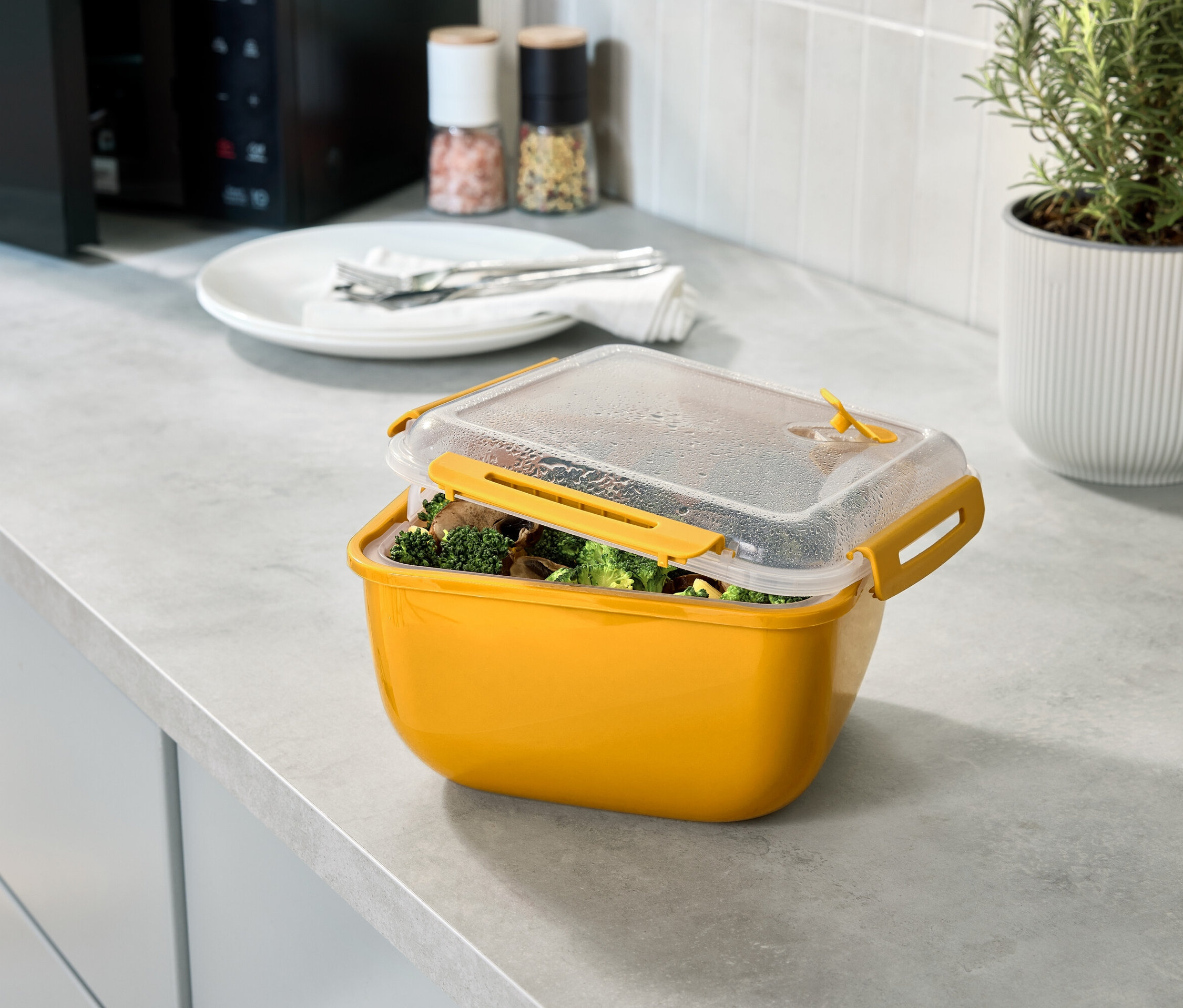Žltá nádoba na potraviny s brokolicou a hubami, s priehľadným vekom, na kuchynskej linke.