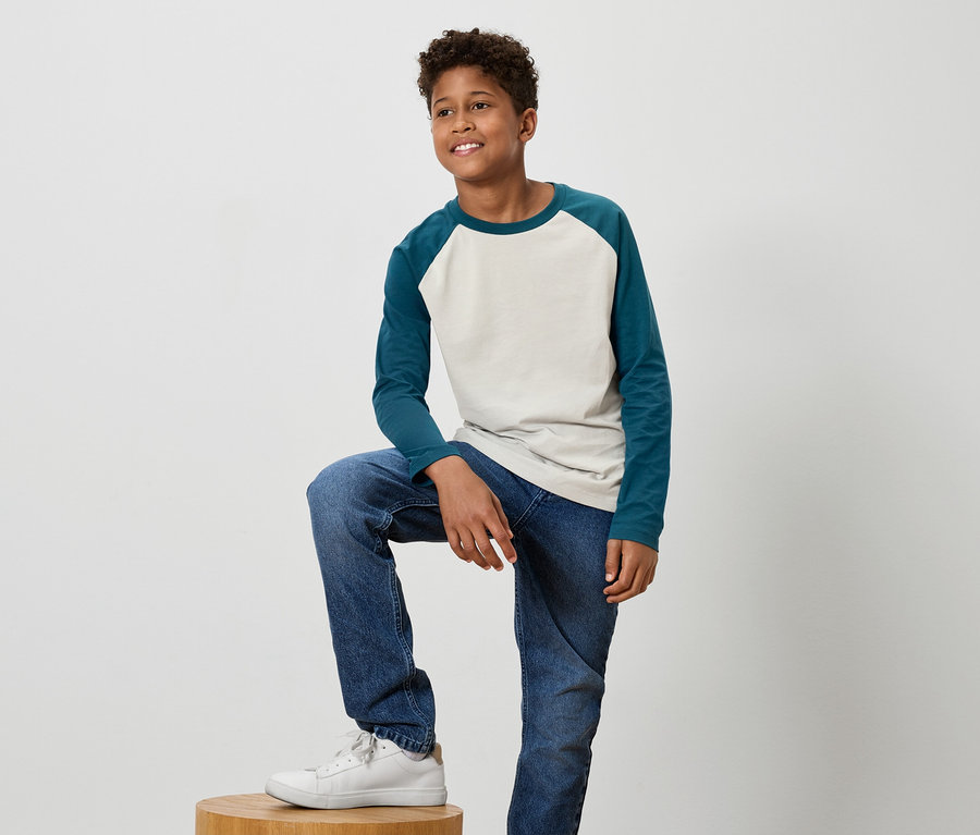 Mladý chlapec s tmavými vlasmi a úsmevom pózuje s nohou na drevenej stoličke, má na sebe modré džínsy a biele tenisky.