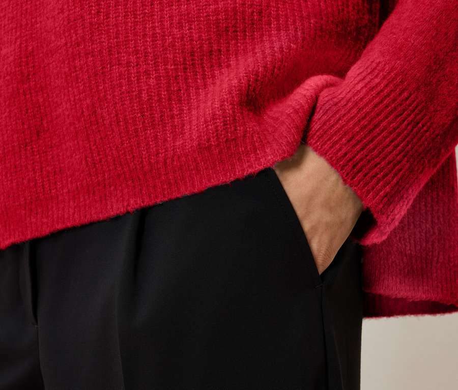 Detail červeného svetra a čiernych nohavíc s rukou vo vrecku.