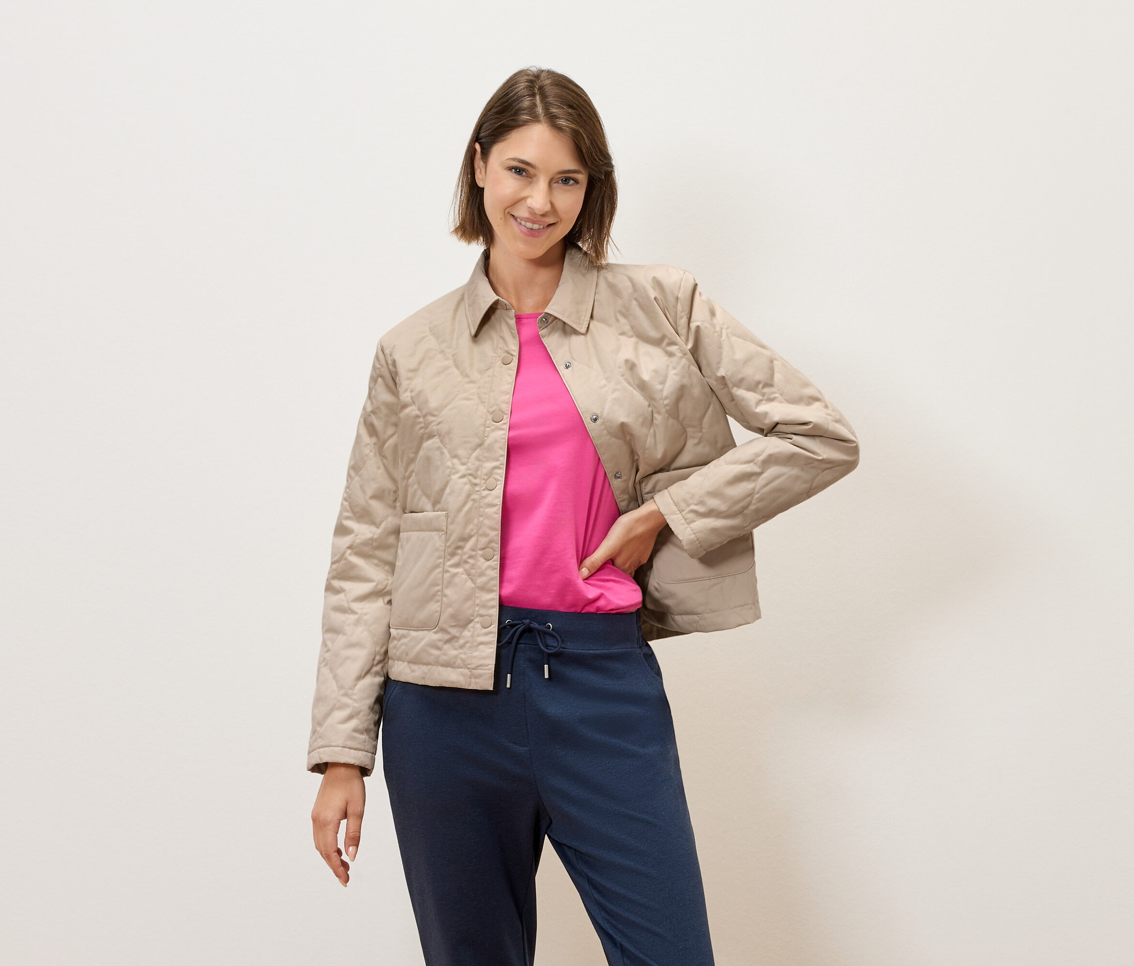 Žena s krátkymi hnedými vlasmi pózuje pred krémovou stenou v béžovej prešívanej bunde, ružovom tričku a modrých nohaviciach.